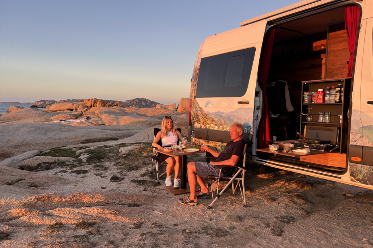 Paar sitzt vor VanEssa Camper am Esstisch bei geoeffnetem Campingsprinter